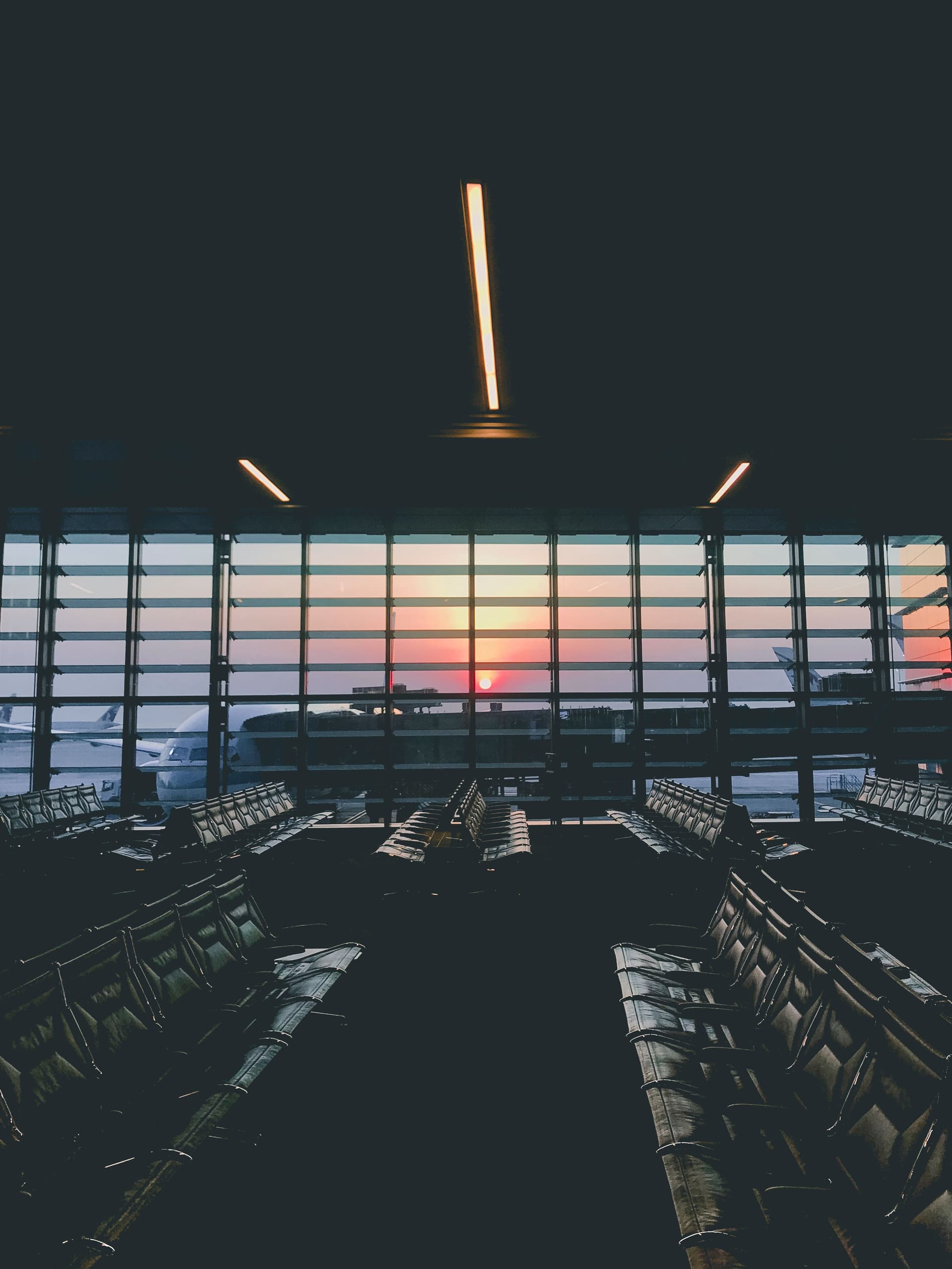 An empty airport terminal at sunset, rows of seats in front of tall windows looking out on parked aircraft.