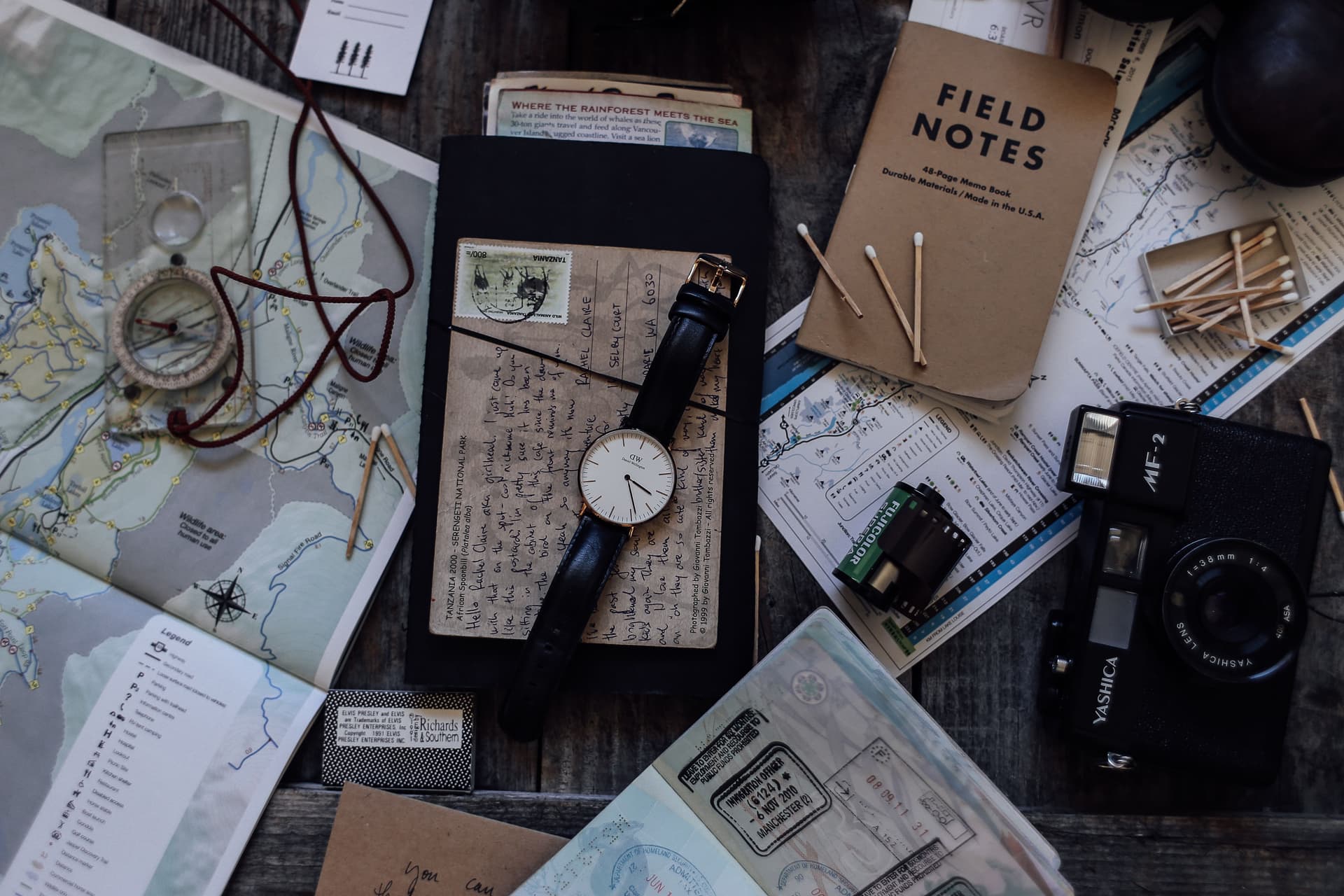 Travel things laid out on a wooden table: folded maps, an old camera and a wristwatch.